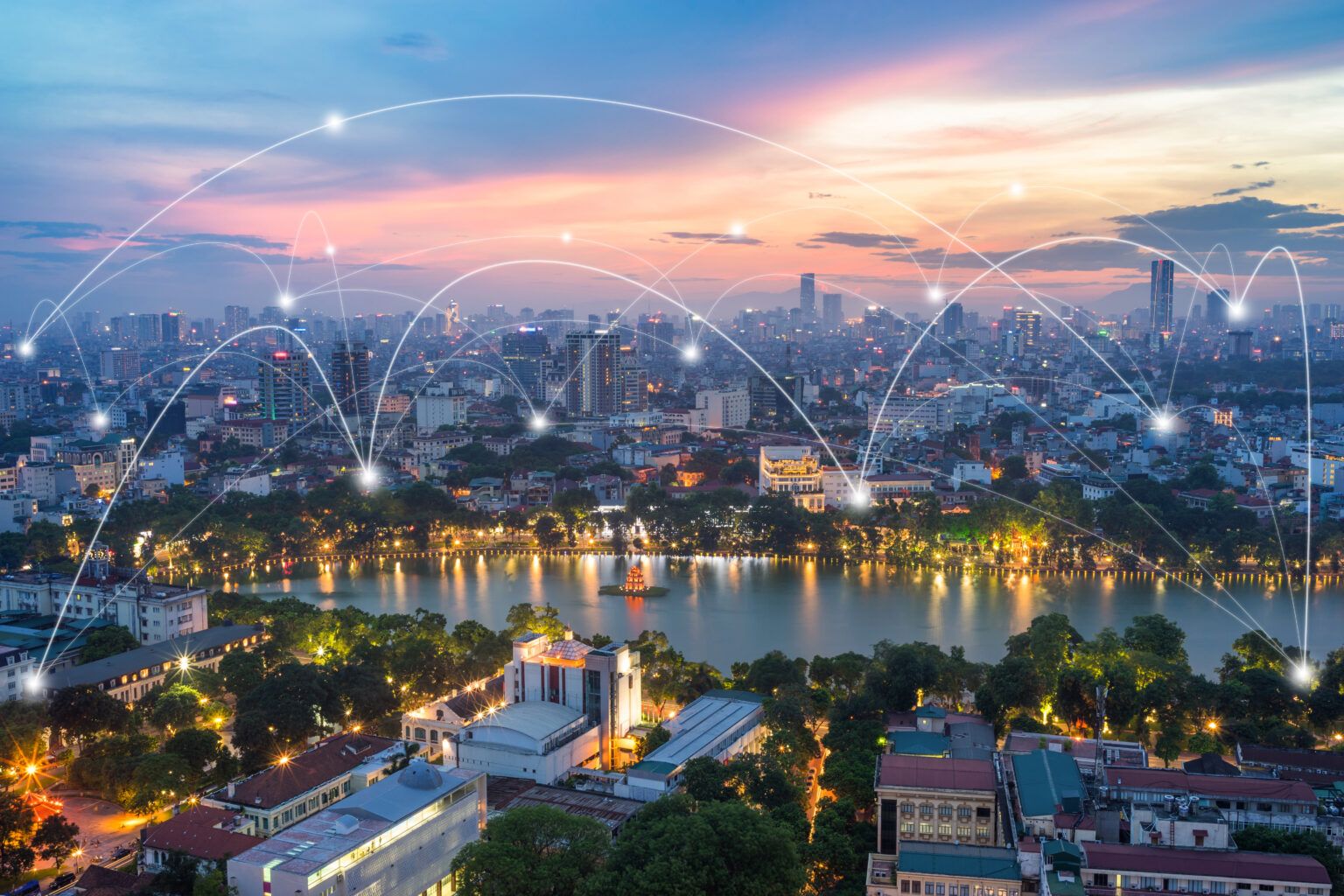 A cityscape at dusk with buildings and a river in the foreground, illuminated by city lights. White lines and glowing points are superimposed over the image, representing digital or network connections across the city.