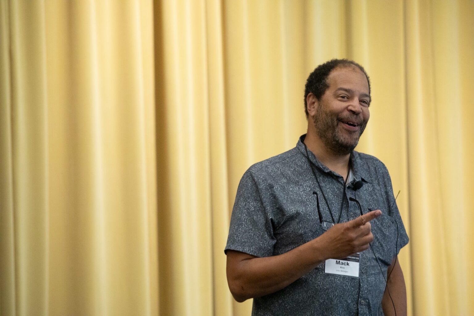 A person smiles in front of a yellow drop curtain, smiling, wearing a collared shirt and a name tag that says "Mack."