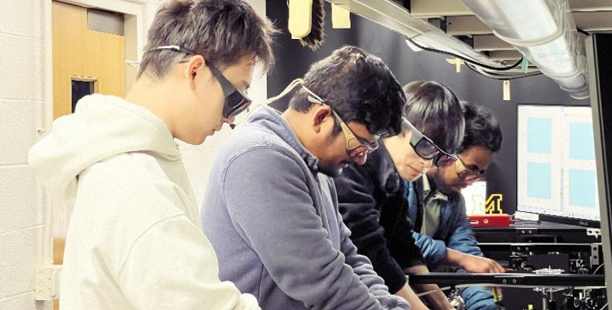 A group of four wearing safety glasses, working with equipment in a lab.